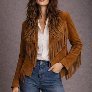 May include: A brown suede jacket with long fringe detailing on the sleeves and chest. The jacket is worn over a white button-down shirt and blue jeans. The jacket has a classic, bohemian style.
