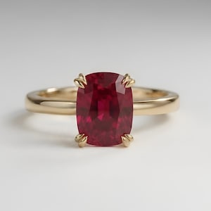 May include: A gold ring featuring a cushion-cut deep red ruby gemstone set in a four-prong setting. The band is smooth and polished, showcasing a simple design that highlights the vibrant color and faceted surface of the central stone.