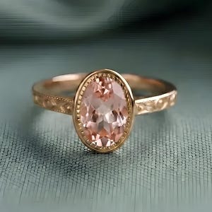 May include: A rose gold ring featuring an oval-cut peach-colored gemstone set in a bezel with a beaded edge. The band displays an intricate engraved pattern along its sides, providing a detailed texture to the metal surface of the jewelry piece.