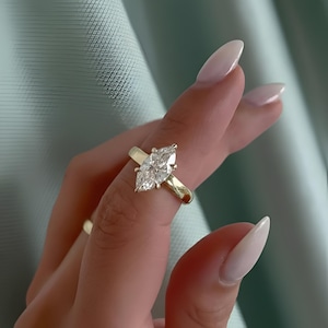 Peut inclure: Une bague en or ornée d'un diamant taille marquise. Le diamant est serti dans un anneau en or. La bague est présentée sur un fond vert clair et doux.
