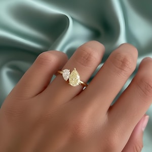 May include: A gold ring with two pear-cut gemstones. One is a clear diamond, the other a yellow diamond. The ring is set on a gold band. The ring is shown on a hand. The gemstones are displayed against a soft, light blue background.