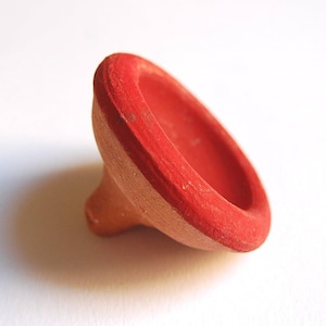 Może przedstawiać: Mały, terakotowy bąk z jasnoczerwoną obwódką. Bąk ma zaokrąglony, miseczkowaty kształt z wąską, stożkową podstawą. Czerwona farba wykazuje pewne zużycie, co dodaje mu rustykalnego uroku.