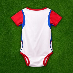 Puede incluir: Un body blanco para beb&eacute; con detalles rojos y azules. Las mangas cortas y los bordes son rojos, con rayas azules en los laterales. Este body c&oacute;modo est&aacute; dise&ntilde;ado para un uso f&aacute;cil.