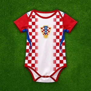 Puede incluir: Un body para bebé con los colores de la selección croata. El body blanco tiene detalles a cuadros rojos y blancos en los laterales y mangas rojas. El escudo de armas croata está centrado en el pecho.