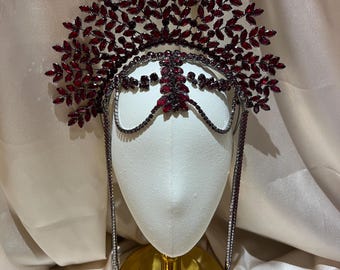 May include: A decorative headpiece featuring a large, fan-shaped crest composed of numerous dark red, leaf-shaped crystals. The piece includes a central forehead ornament and two long, dangling crystal-encrusted chains that frame the sides.