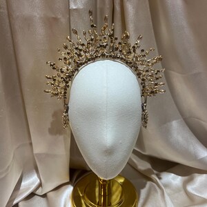 May include: A gold-toned crown headpiece featuring an intricate arrangement of numerous light brown and clear crystals. The headpiece is constructed with a thin metal band and radiates upward with multiple crystal-tipped branches of varying heights.