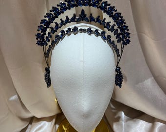 May include: A double-tiered crown featuring dark blue marquise-cut crystals arranged in a leaf-like pattern. The headpiece is constructed with a dark metal frame, showcasing a tall, arched design with smaller crystal accents at the base sides.