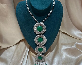 May include: A silver-toned necklace featuring a chain and four vertically aligned circular floral-shaped pendants, each centred with a smooth green stone. The bottom pendant includes dangling silver-toned charms and small green accents.