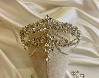 May include: A silver-toned tiara with numerous clear crystals. The crown features a central, pointed crystal and a floral-shaped pendant. The tiara is displayed on a neutral-colored head form against a backdrop of white satin fabric.