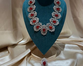 May include: A jewellery set consisting of a necklace, a pair of earrings, a ring, and a bracelet. Each piece features an intricate silver-toned filigree metal frame surrounding a smooth, oval-shaped red stone set in the centre of each decorative link.