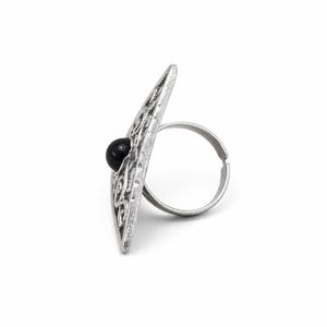May include: A silver-toned ring featuring a large, elongated, diamond-shaped face with an intricate openwork scroll pattern and a single, smooth, round black bead set in the centre. The band is thin, circular, and has an adjustable open gap.