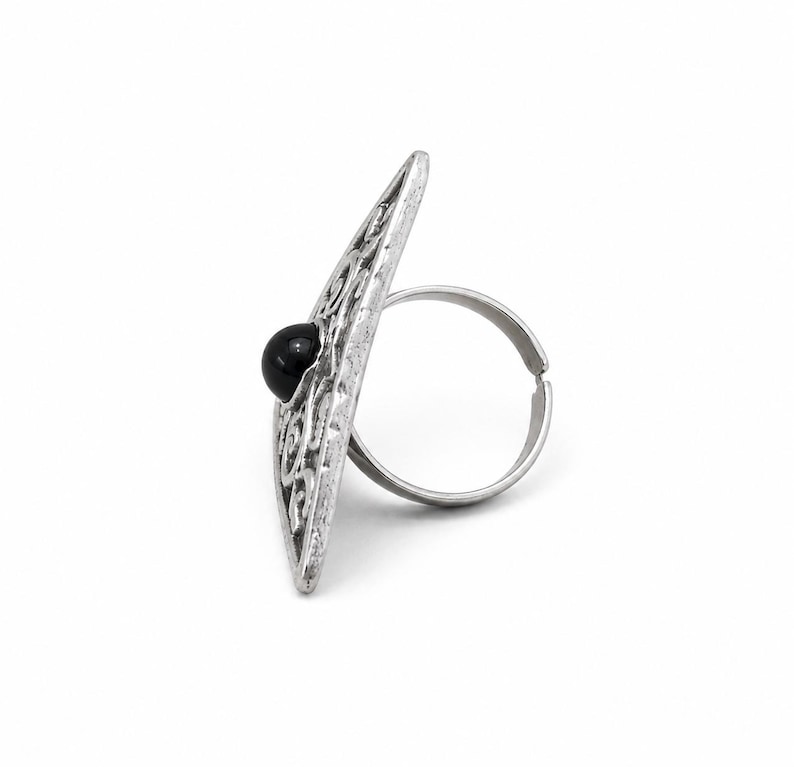 May include: A silver-toned ring featuring a large, elongated, diamond-shaped face with an intricate openwork scroll pattern and a single, smooth, round black bead set in the centre. The band is thin, circular, and has an adjustable open gap.