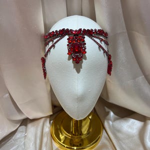 May include: A headpiece featuring deep red faceted crystals set in a dark metal frame. The design includes a central ornate cluster of marquise and oval-shaped stones, with delicate chains of smaller red crystals draping along the sides.