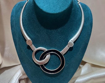 May include: A silver-toned metal collar necklace featuring a rigid curved bar design that connects to two interlocking circular pendants. One circle is solid silver-toned metal, while the other is a silver-toned ring with a black inlay centre.