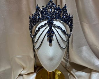 May include: A dark blue crystal crown featuring an intricate metal frame adorned with numerous faceted navy blue stones. The headpiece includes delicate dangling chains that drape across the sides and a central teardrop-shaped pendant.