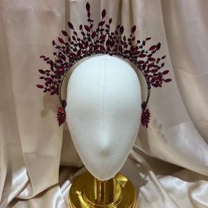May include: A crown-style headpiece featuring a dark metal frame adorned with numerous deep red, marquise-cut crystals. The crystals are arranged in a radiating, spiky pattern across the top, with additional crystal clusters at the sides.