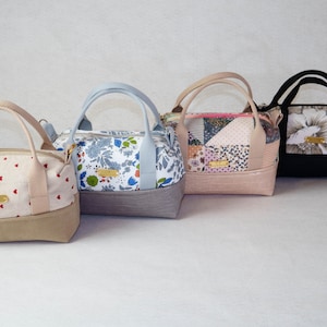 Könnte beinhalten: Gezeigt werden vier kleine, farbenfrohe Handtaschen mit Tragegriffen. Die Taschen haben verschiedene Muster, darunter Blumen-, Patchwork- und Herzdesigns. Jede Tasche hat einen kontrastierenden Boden und ein kleines goldfarbenes Etikett.