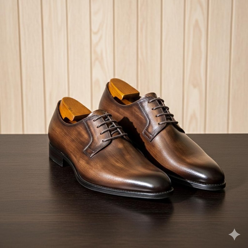 May include: A pair of brown leather Oxford shoes with wooden shoe trees. The shoes have a classic design with laces and a polished finish. They are displayed on a dark surface against a light wood panel background.