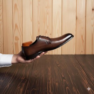 May include: A brown leather dress shoe with a classic design, held in a person's hand. The shoe has a polished finish, laces, and a wooden shoe tree inside. The background features a wooden wall and table.