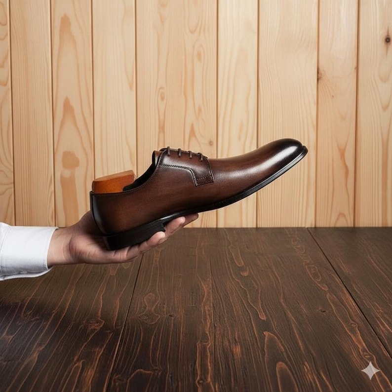 May include: A brown leather dress shoe with a classic design, held in a person's hand. The shoe has a polished finish, laces, and a wooden shoe tree inside. The background features a wooden wall and table.