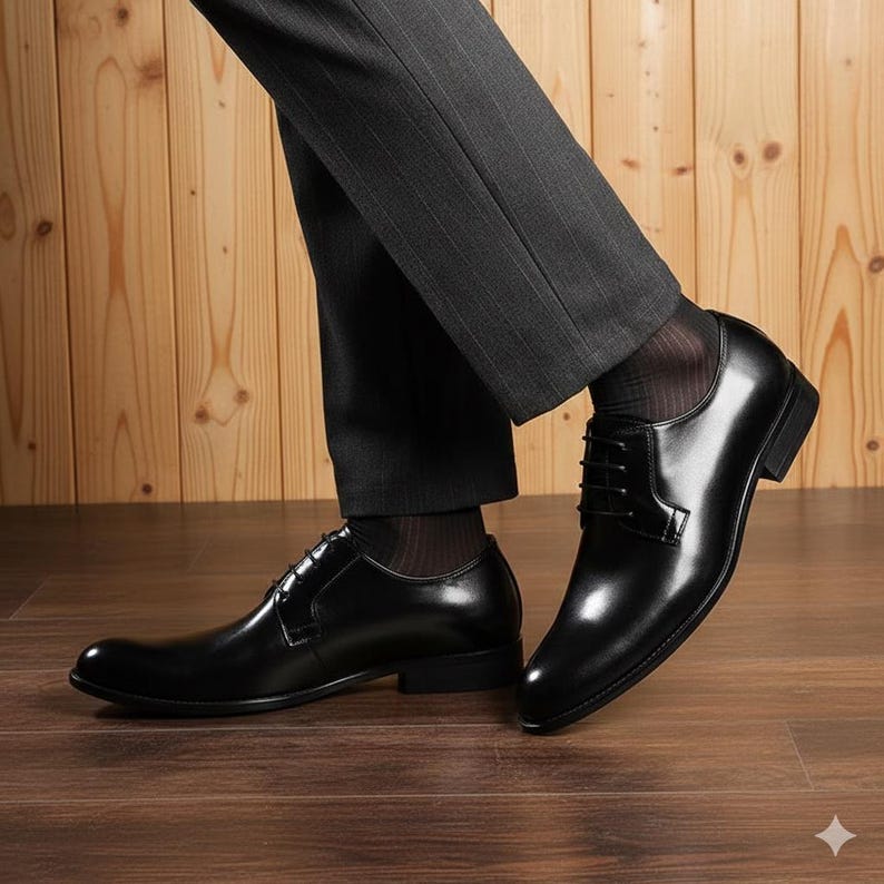May include: A pair of black leather dress shoes with a glossy finish. The shoes have a classic lace-up design and are worn with dark grey dress trousers and black socks. The shoes are on a wooden floor.