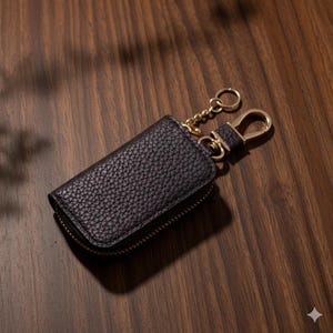 May include: A black leather key case with a gold-colored zipper and hardware. The key case has a textured surface and a gold chain and clasp for attaching keys. The case is closed and sits on a dark wooden surface.