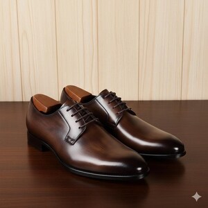 May include: A pair of brown leather dress shoes with dark brown laces. The shoes have a polished finish and are displayed on a dark wooden surface. Shoe trees are inserted in the shoes.