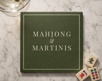 Op de afbeelding: Een vierkante, donkergroene onderzetter met de woorden "MAHJONG & MARTINIS" in witte letters. De onderzetter ligt op een marmeren oppervlak, met drie mahjongtegels en een martini glas met olijven.