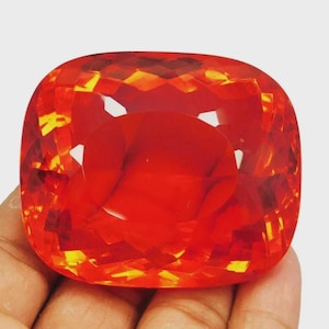 May include: A large, faceted gemstone with a vibrant orange-red hue. The cushion-cut stone displays a complex pattern of light refraction, creating a sparkling effect. The gem is held in a hand, showcasing its size and color.