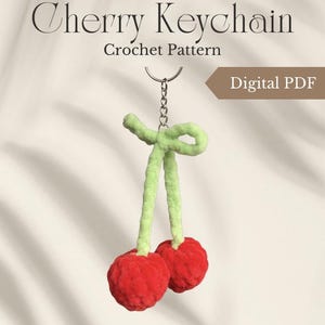 Könnte beinhalten: Ein gehäkelter Kirsch-Schlüsselanhänger mit roten Kirschen und grünen Stielen. Der Schlüsselanhänger ist an einer silbernen Kette befestigt. Der Text "Cherry Keychain Crochet Pattern" befindet sich oben im Bild, mit "Digital PDF" in einem Banner.