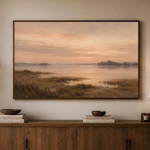 Könnte beinhalten: Ein rechteckiger Wandkunstdruck, der einen ruhigen See bei Sonnenuntergang in sanften Pfirsich- und Goldtönen zeigt, gerahmt in einem dunklen Holzrahmen. Das Bild zeigt ruhiges Wasser, ferne Bäume und sumpfige Gräser unter einem dunstigen Himmel.