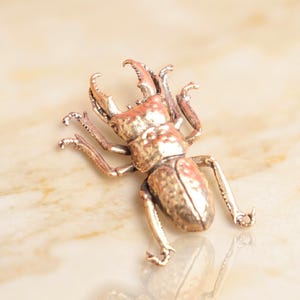 Puede incluir: Figurita detallada de escarabajo plateado, con mandíbulas prominentes y detalles intrincados en las patas. El cuerpo del escarabajo tiene una superficie texturizada y el diseño es realista. La figura se encuentra sobre un fondo claro, similar al mármol.