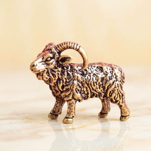 May include: A small, metallic bronze-toned figurine of a ram standing in profile. The sculpture features detailed, textured fur across its body, a curved horn extending from its head, and four legs with defined hooves, capturing the animal's form.