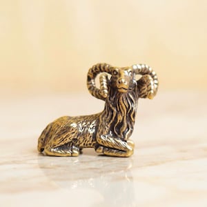 May include: A small, metallic gold-colored figurine of a ram in a resting position. The sculpture features detailed, textured fur, prominent curved horns, and a focused facial expression, crafted with a polished finish that reflects light.