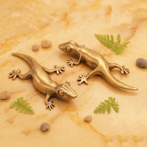 Puede incluir: Dos figuras de lagarto de color bronce con detalles precisos, colocadas sobre una superficie clara y estampada. Pequeños guijarros y hojas verdes de helecho están dispersos alrededor de los lagartos, realzando la estética natural. Son objetos decorativos.