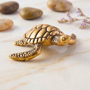 Puede incluir: Una detallada figura de tortuga dorada con caparazón y aletas texturizadas. La tortuga está colocada sobre una superficie similar al mármol, con varias piedras lisas y ramitas de lavanda en el fondo. La figura es un objeto decorativo.