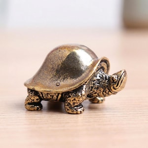 May include: A small, detailed brass turtle figurine. The turtle has a textured shell and legs, with a smooth, polished head. The figurine is a decorative item, likely for display.