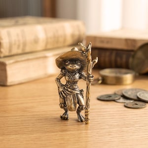 May include: A bronze frog figurine wearing a hat and holding a staff. The frog is dressed in a detailed outfit, with a belt and a skirt. The figurine is standing on a wooden surface, with books and coins in the background.