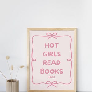 Könnte beinhalten: Ein gerahmter Druck mit dem Text "HOT GIRLS READ BOOKS (duh)" in Rosa auf hellrosa Hintergrund. Der Text ist von einem rosa Zierrahmen mit Schleifen oben und unten umgeben. Der Rahmen besteht aus hellem Holz. Links steht eine kleine Vase mit Trockenblumen.
