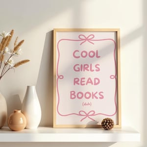 Könnte beinhalten: Ein rechteckiger Wandkunstdruck in einem hellen Holzrahmen mit einem rosafarbenen Zierrahmen, der oben und unten mit Schleifen verziert ist. Der rosafarbene Text lautet COOL GIRLS READ BOOKS (duh) auf hellcremefarbenem Hintergrund.