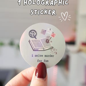 Puede incluir: Una pegatina hologr&aacute;fica redonda que muestra un libro abierto con una huella de mano oscura, una lupa y peque&ntilde;as notas fijadas. El texto dice i solve murder for fun. La pegatina tiene un acabado iridiscente claro y una forma circular.