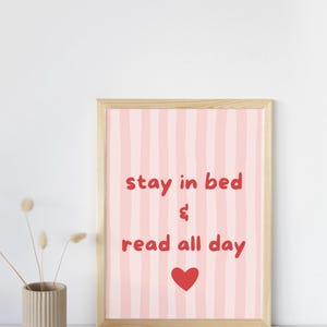 Könnte beinhalten: Ein gerahmter Druck mit einem rosa gestreiften Hintergrund und den roten Worten "stay in bed & read all day", zusammen mit einem roten Herzen. Der Rahmen ist aus hellem Holz. Links steht eine Vase mit Trockenblumen.