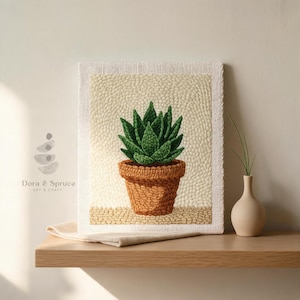 May include: A framed textile art piece featuring a green succulent plant in a brown pot. The artwork is displayed on a wooden shelf with a neutral background. The piece is labeled "Dora & Spruce ART & CRAFT".