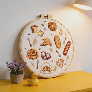 May include: A round embroidery hoop featuring various baked goods, including bread, croissants, and pretzels, in shades of brown and beige. The embroidery is set against a cream-colored fabric, with a wooden hoop. A small potted plant and a yellow duck figurine are in the foreground.