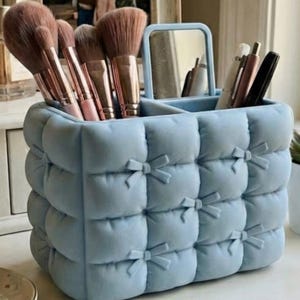 Op de afbeelding: Een lichtblauwe, gewatteerde make-up organizer met strikdetails. Het heeft meerdere compartimenten en een kleine spiegel. De organizer bevat make-up borstels en andere beauty tools.