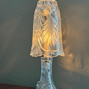 May include: A clear glass table lamp with a decorative, etched floral design. The lamp has a flared base, a slender stem, and a bell-shaped shade. The shade is etched with a leaf pattern. The lamp is lit, casting a warm glow.