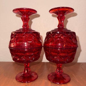Puede incluir: Dos copas vintage de color rojo rubí. Cada copa presenta una base redonda, un tallo, un cuenco con diseño de diamantes y un borde acampanado. Las copas son de vidrio y se exhiben sobre una superficie de madera.