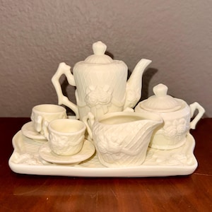 May include: A cream-coloured miniature tea set, including a teapot, sugar bowl, creamer, two teacups, and saucers, all arranged on a rectangular tray. The set features a textured, embossed design with floral accents.