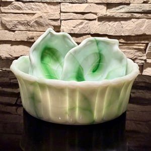 Op de afbeelding: Een decoratieve kom in vintage-stijl met twee blad-vormige schalen erin. De kom en schalen zijn crèmewit met groene marmering. De kom heeft een gegroefd ontwerp aan de zijkanten. De achtergrond is een bakstenen muur.