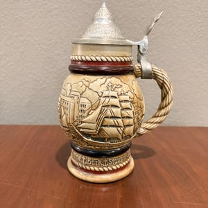 May include: A beige ceramic beer stein with a detailed relief of a sailing ship and buildings. It has a metal lid and a twisted handle. The base reads "The Bark Staysail".